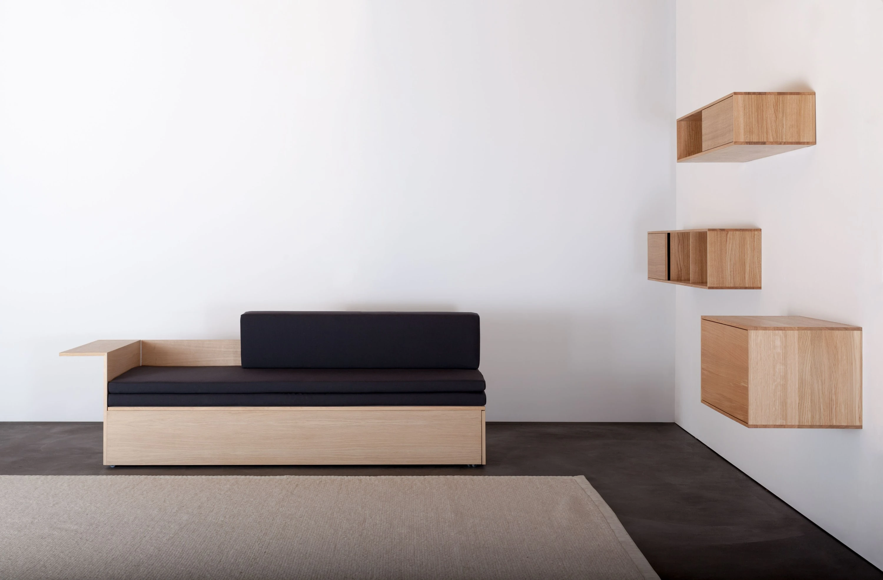 Couch aus Holz mit schwarzer Auflage auf hellem Teppich, rechts drei Wandregale aus Holz.