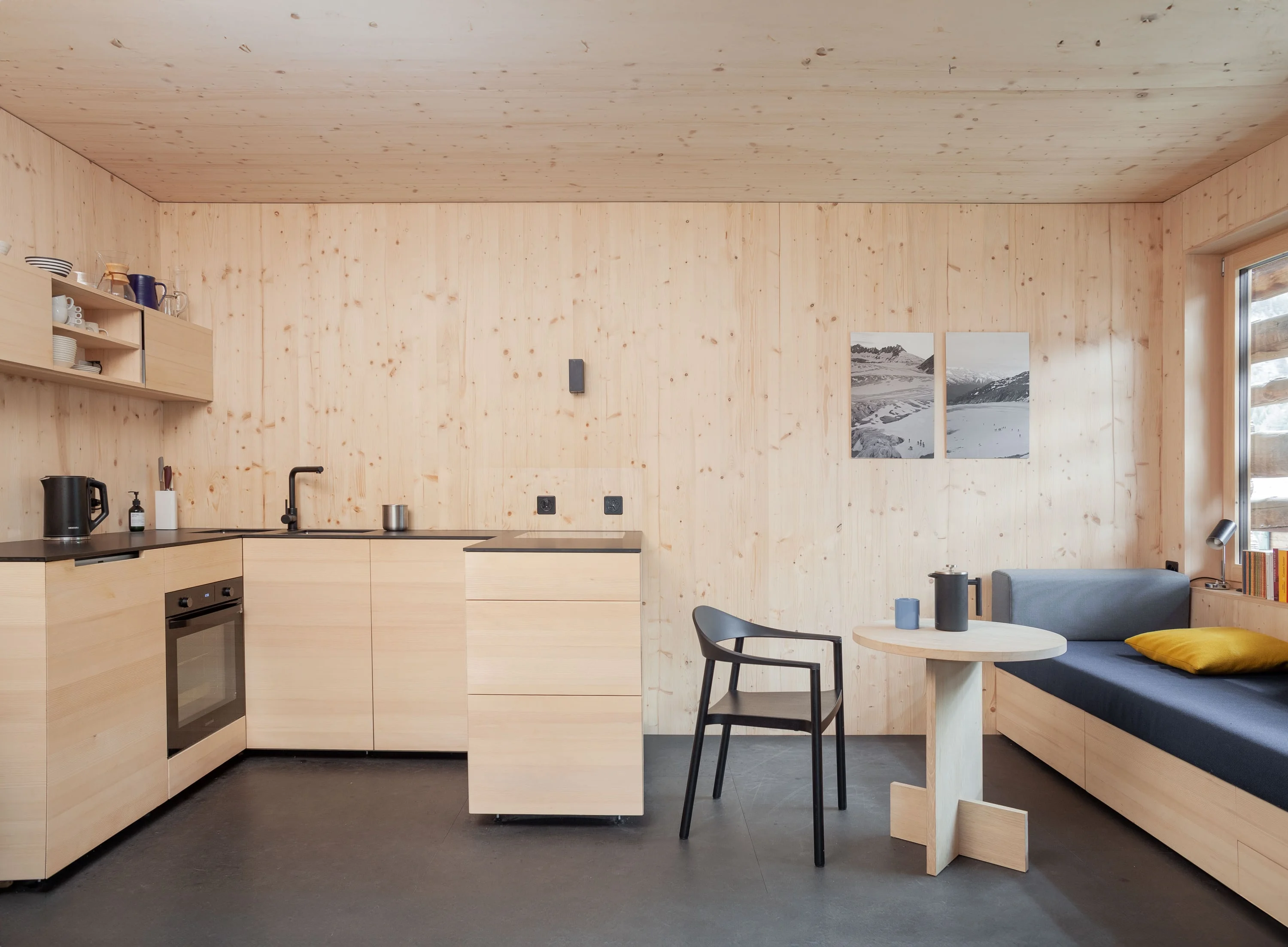 Wohnküche aus hellem Holz mit Küchenzeile, kleinem Tisch und Sitzbank mit gelben Kissen.