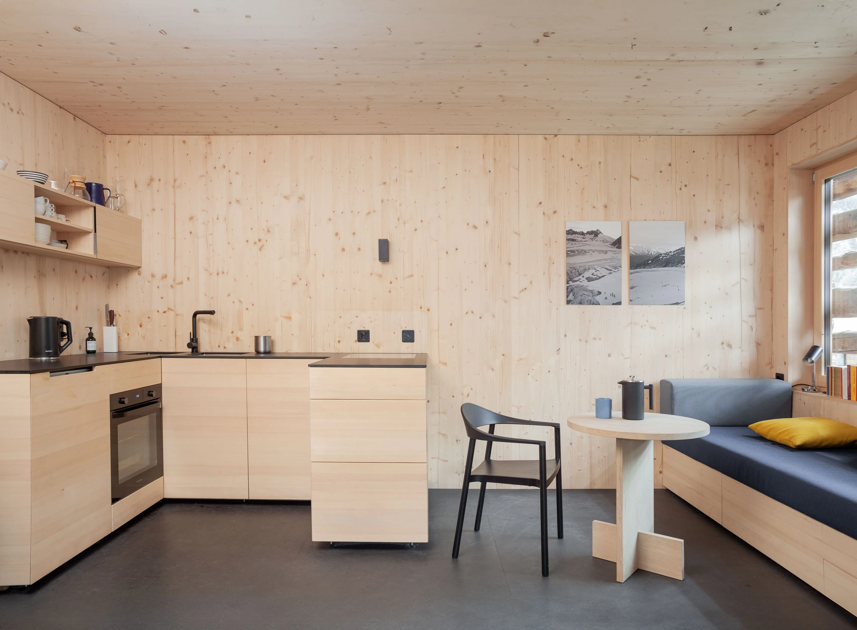 Wohnküche aus hellem Holz mit Küchenzeile, kleinem Tisch und Sitzbank mit gelben Kissen.