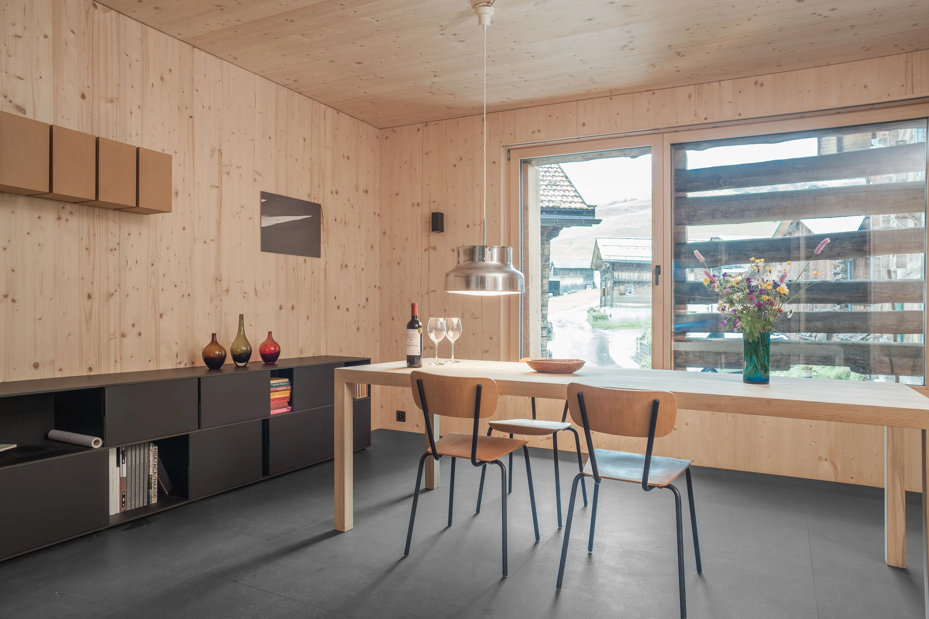 Raum aus hellem Holz mit schwarzem Sideboard, Esstisch und Fenster vor den Rundhölzern des Stalls.