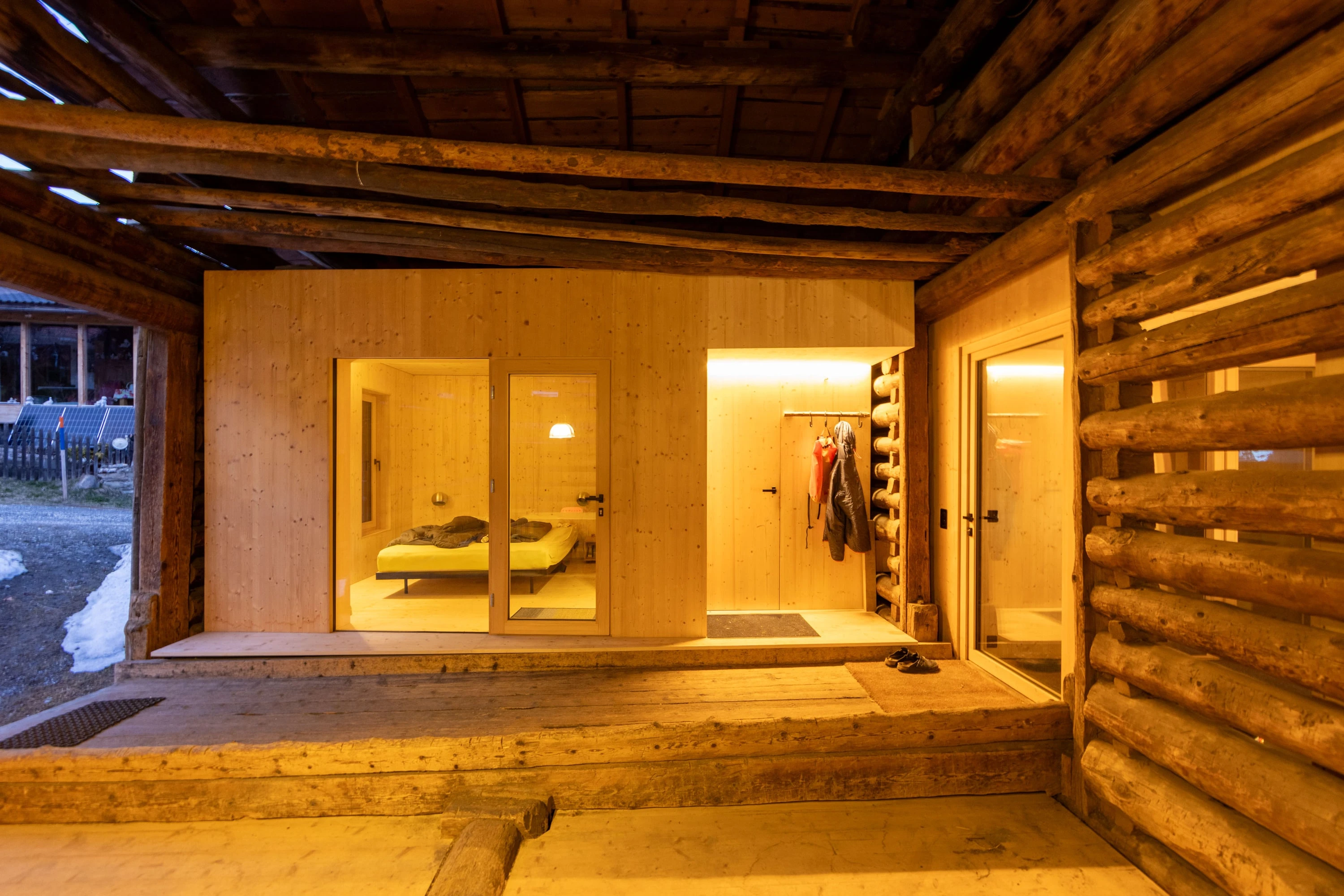 Blick aus dem dunklen Stallraum auf den beleuchteten Holzkubus mit Schlafzimmer und Badezimmertür.
