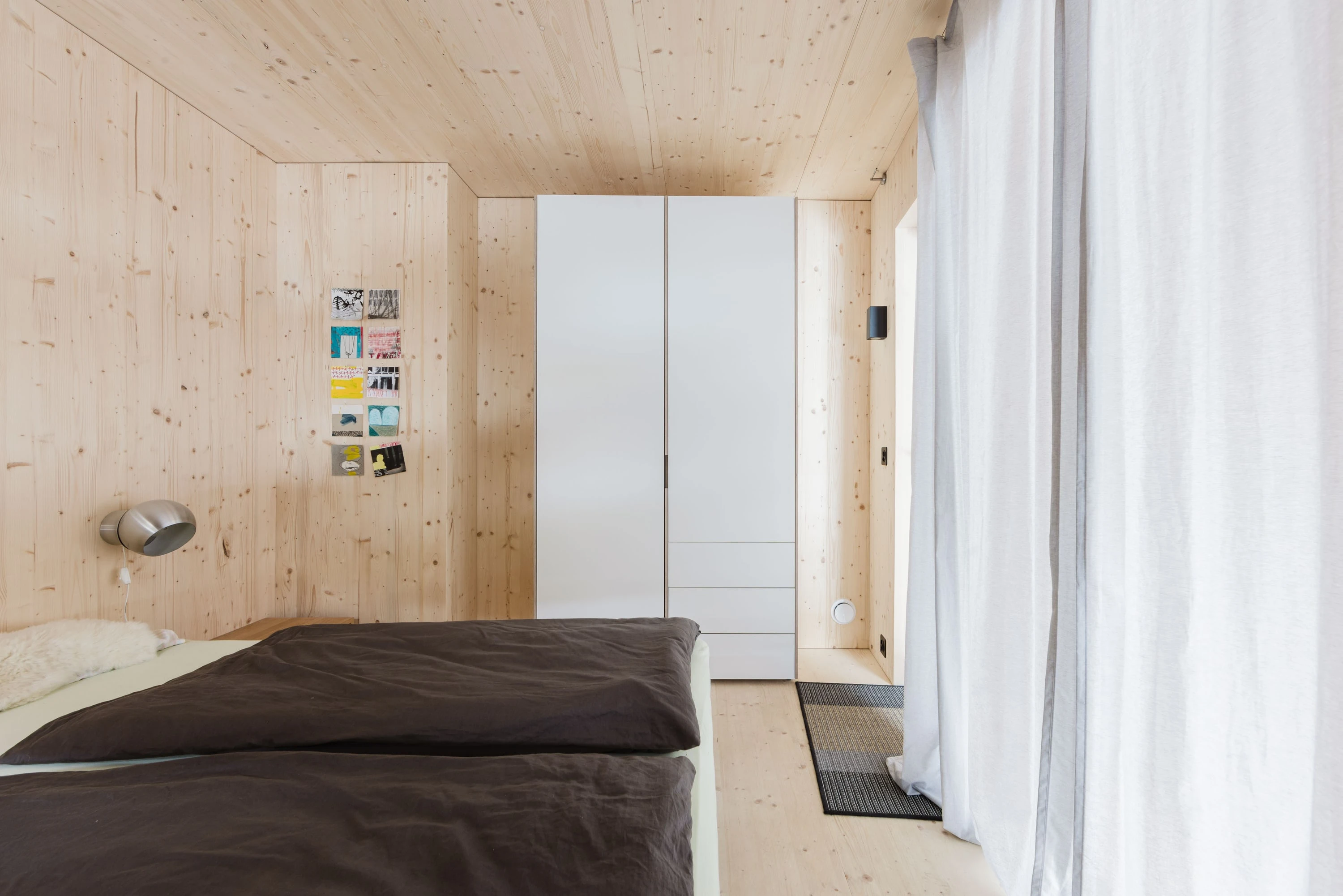 Schlafzimmer aus hellem Holz mit Bett, weissem Schrank und Vorhang.