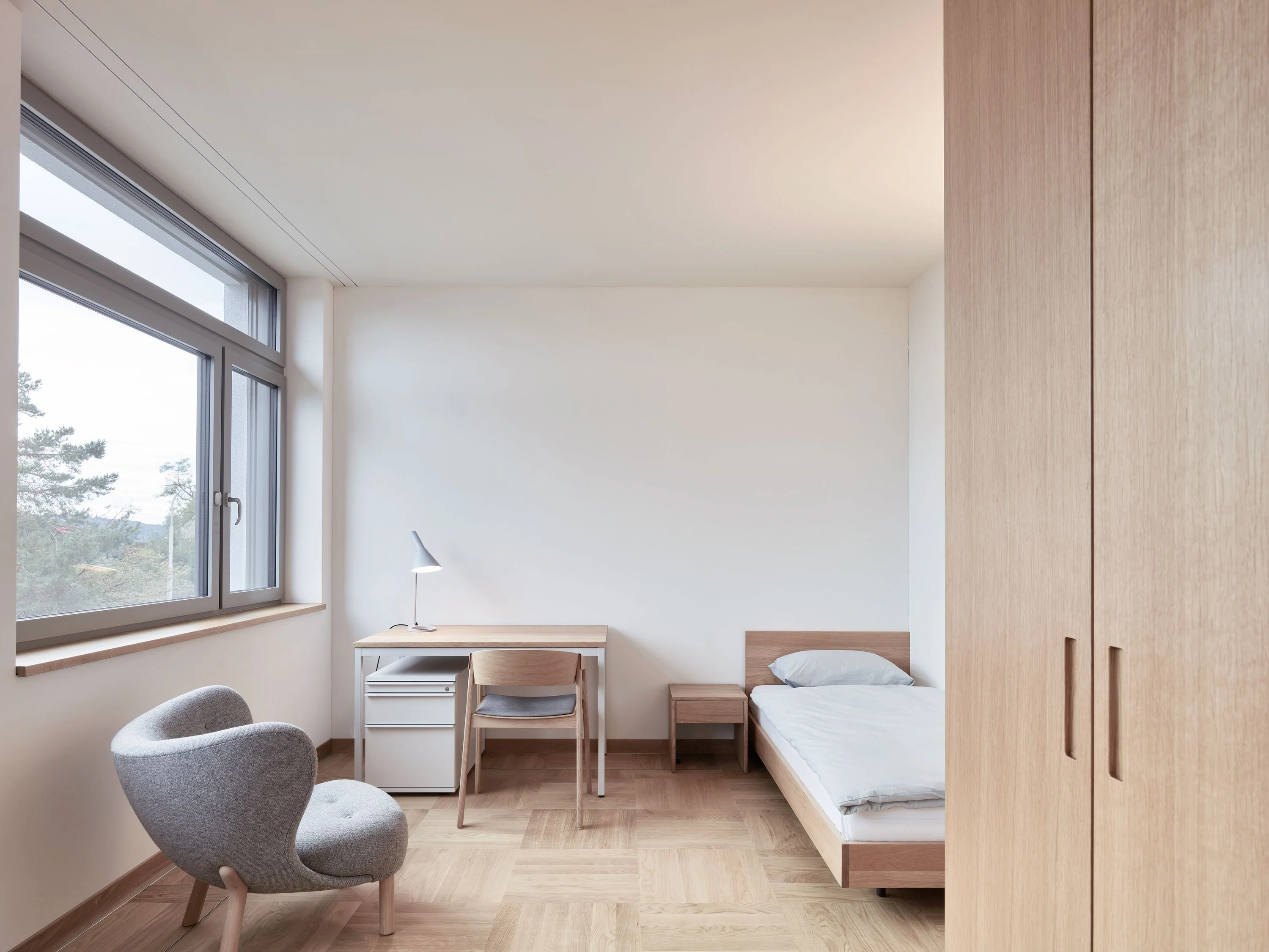 Patientenzimmer mit Bett, Sessel, Schreibtisch und Holzschrank.