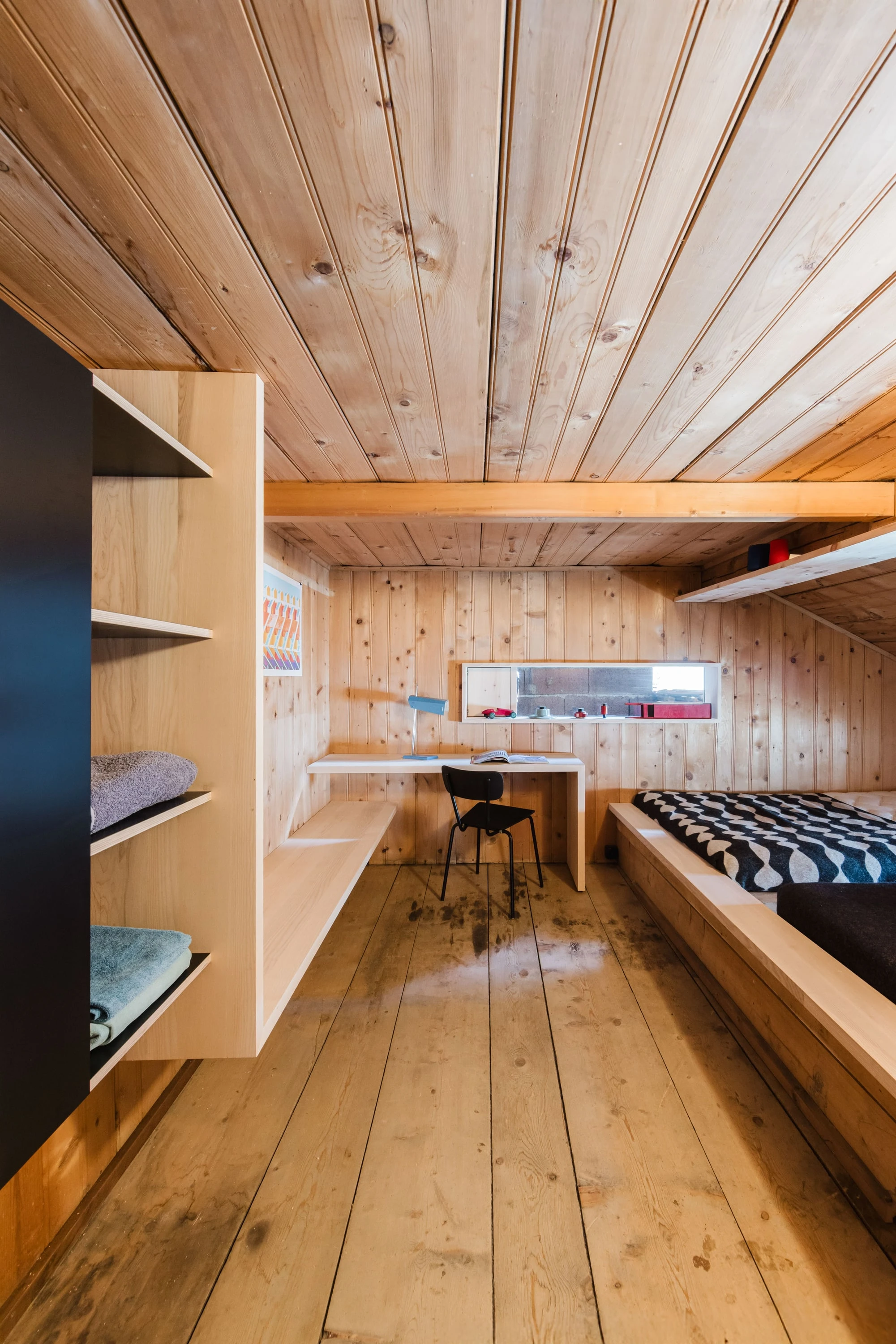 Kleines Dachzimmer aus Holz mit Regal, Schreibtisch und Schlafpodest.