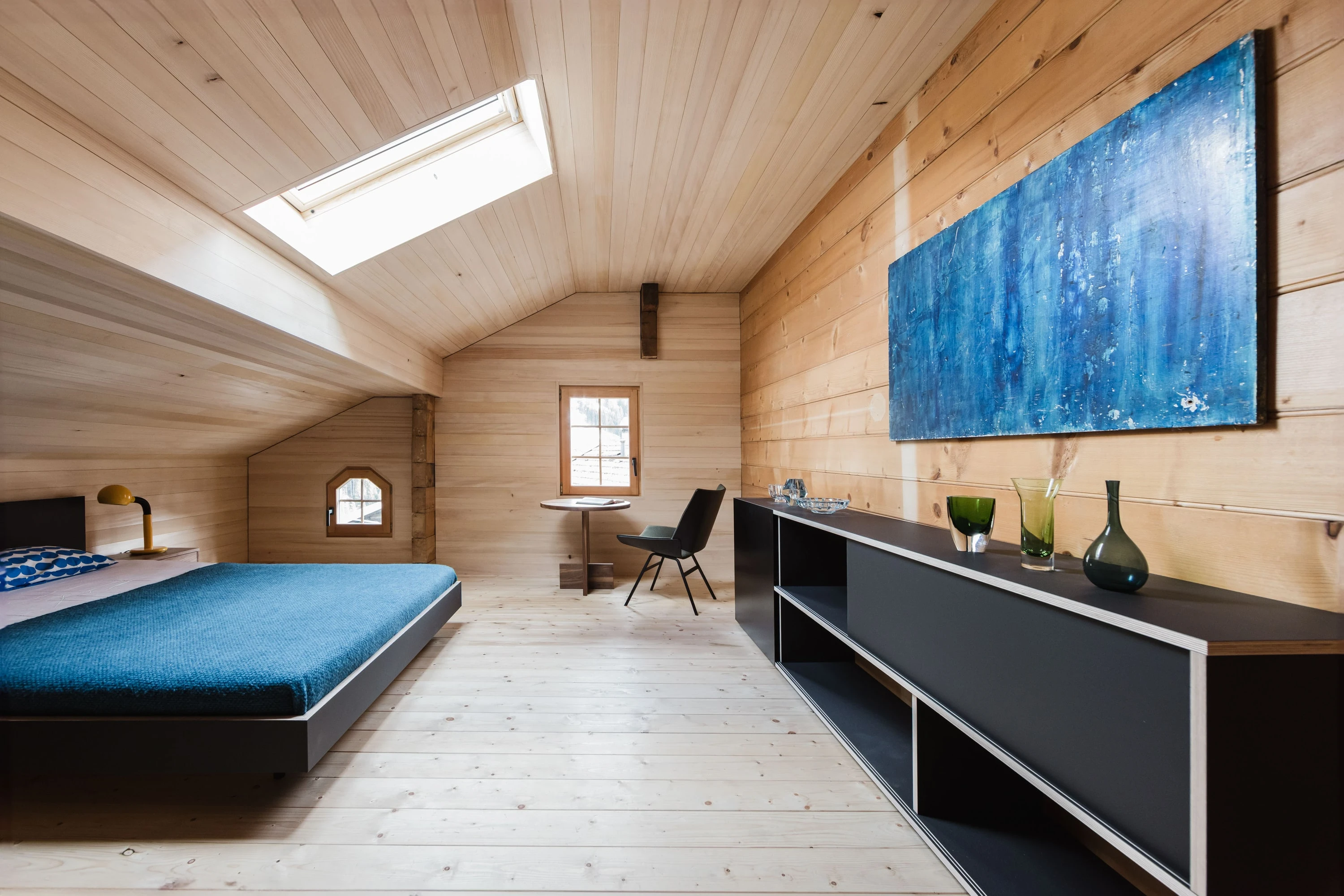 Grosses Dachzimmer mit blauem Bett, schwarzem Lowboard und grossem blauem Bild.