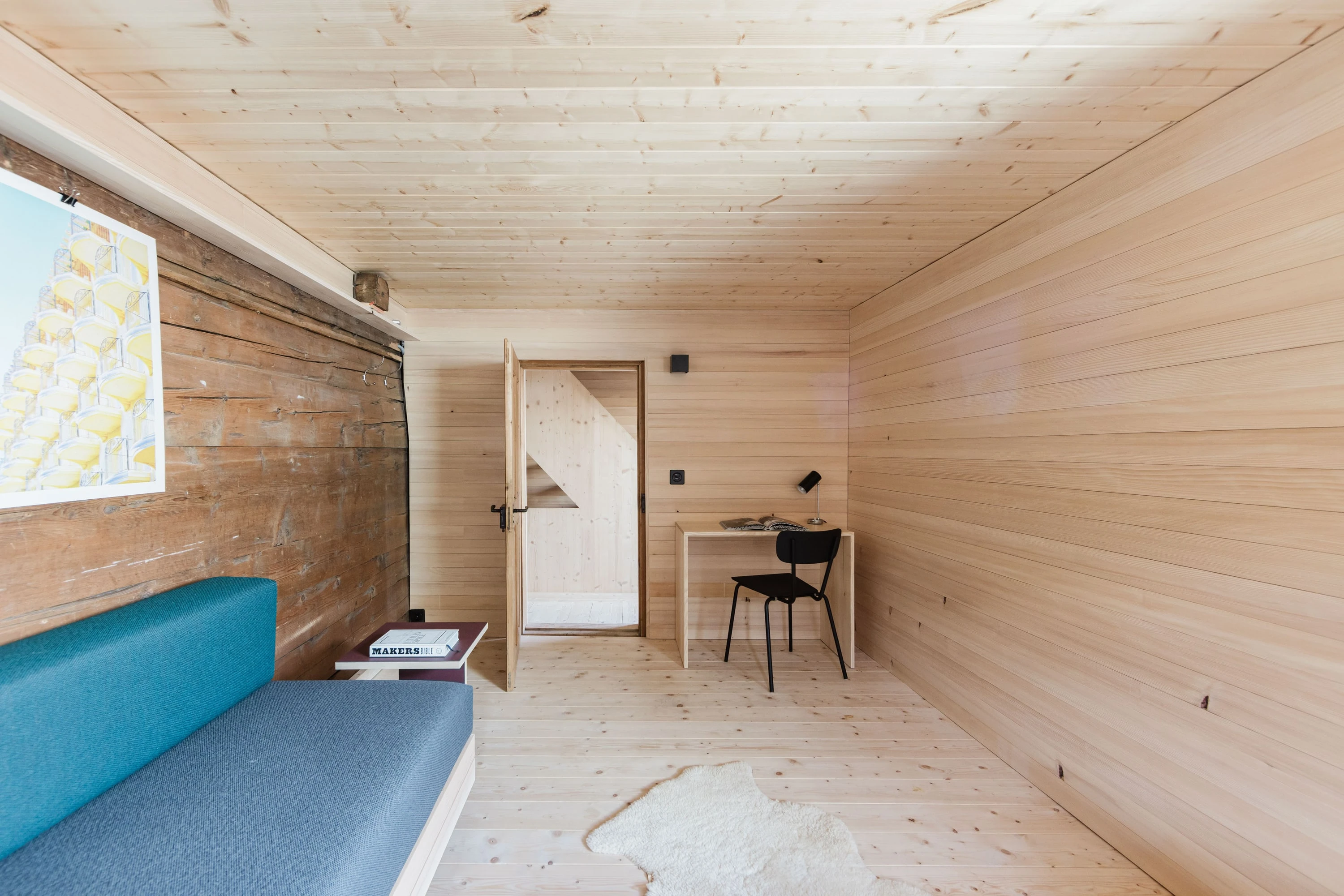 Zimmer mit Holzwänden, blauer Sitzbank und kleinem Schreibtisch neben der Tür.