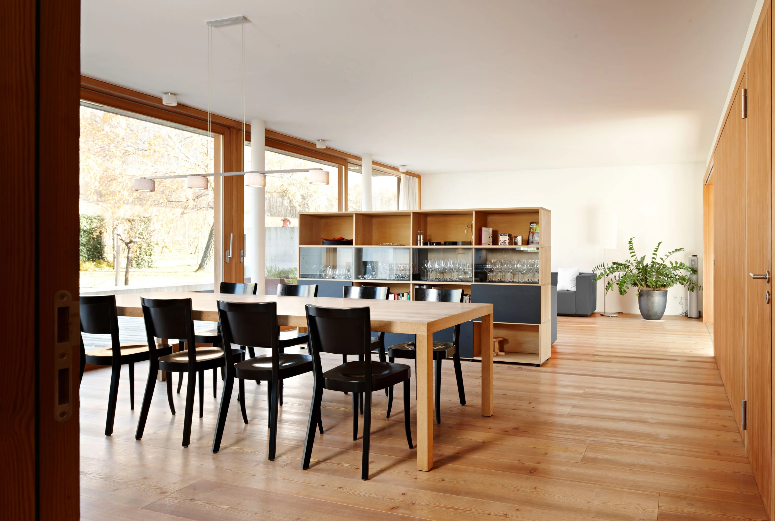 Essraum mit langem Holztisch, schwarzen Stühlen und Regal mit Gläsern vor der Fensterfront.