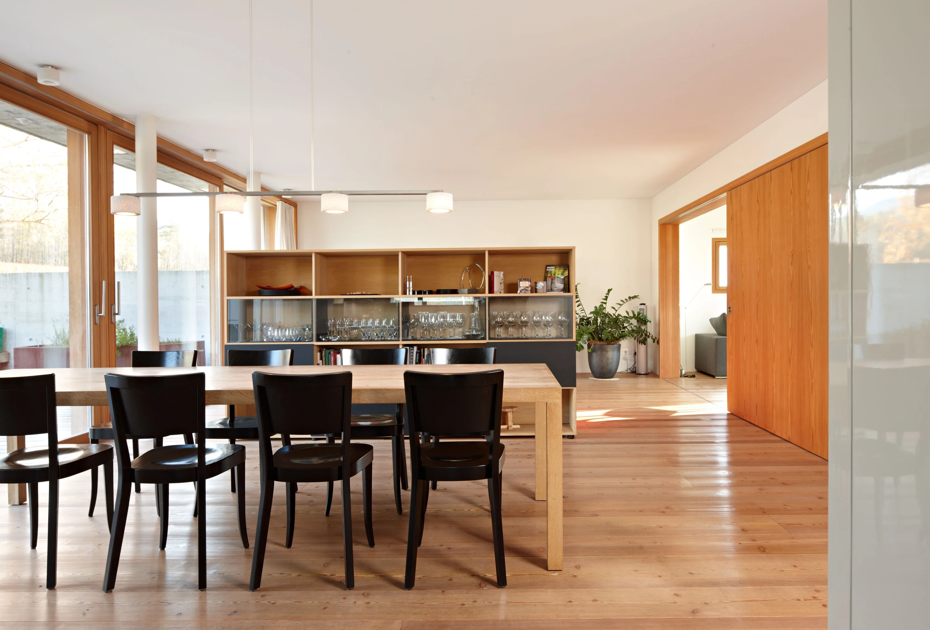 Essraum mit Holztisch, schwarzen Stühlen, Regal und grossen Fenstern.